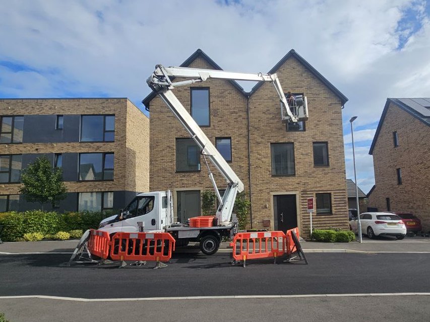 Truck-mounted cherry picker with the basket at roof level on a new-build housing development, barriers set out around the vehicle